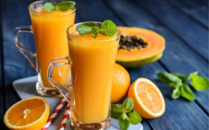 Mamão parido ao meio ao fundo, e na frente dois copos de bebida da mesma fruta.